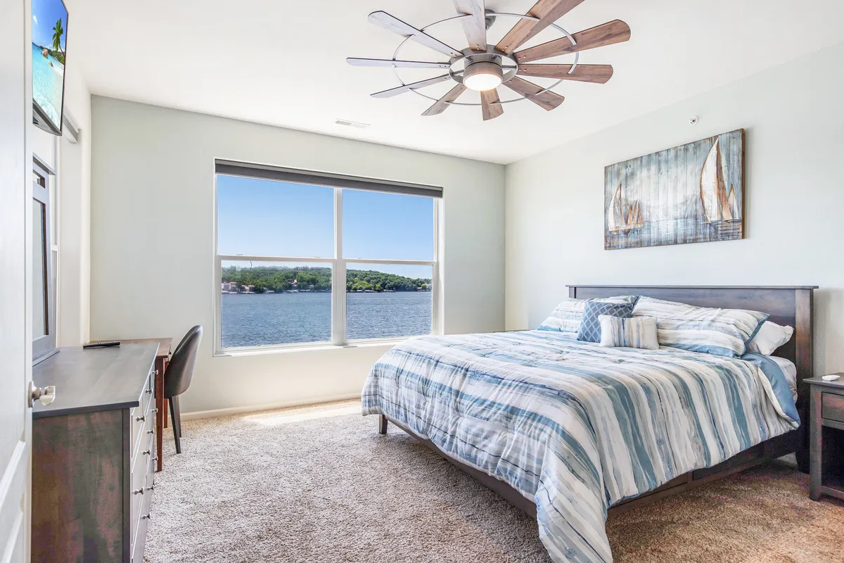 A serene bedroom featuring a king-sized bed with striped bedding, large window looking out over the water, and tasteful wall art. The decor is calm and welcoming, inviting relaxation.
