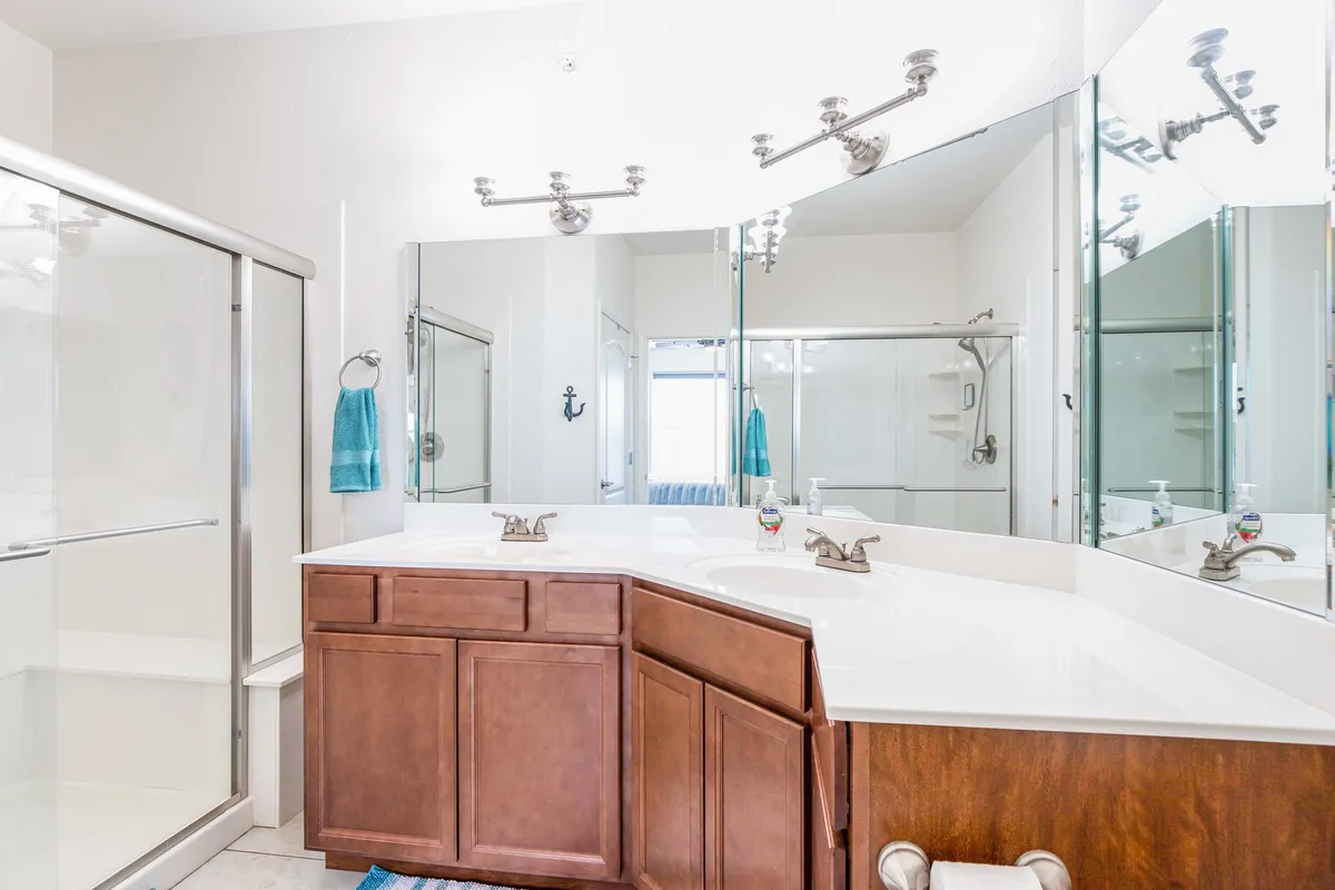 A bright, modern bathroom with a double vanity sink, large mirrors, a spacious shower area, and blue towel accents. Sunlight pours in, creating an inviting space for relaxation and self-care.