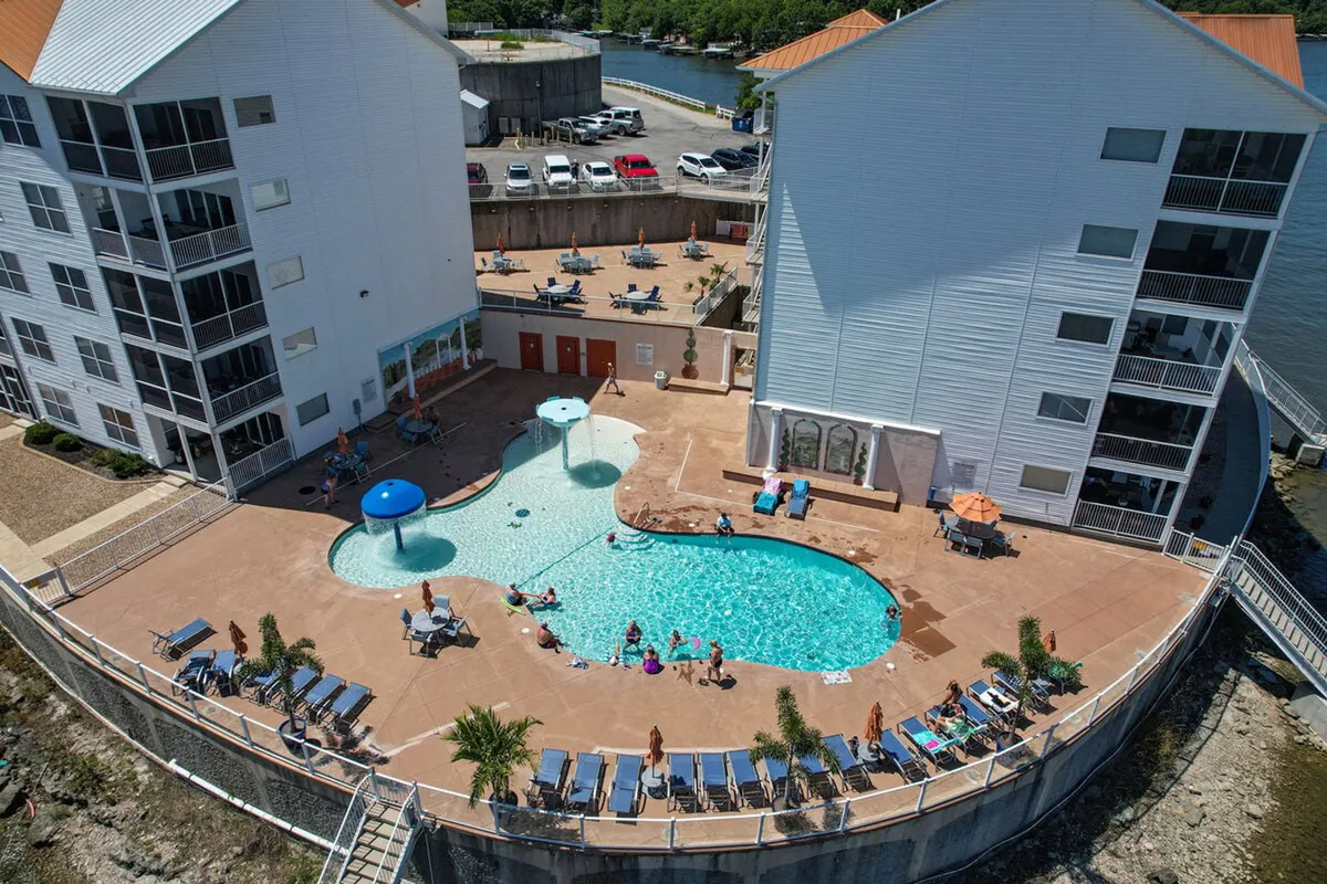 A vibrant outdoor scene featuring a sparkling swimming pool surrounded by lounge chairs and water features, with the condo buildings in the background. The pool area is spacious, inviting, and clearly designed for both kids and adults to enjoy recreational activities together.
