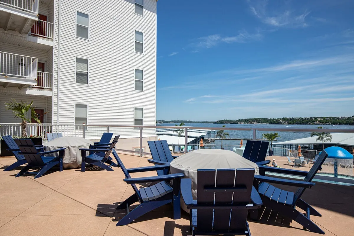 This exterior image shows blue Adirondack chairs arranged on a patio or balcony overlooking a serene expanse of lake water, with the condo building's white facade partially visible. The setting suggests a relaxed, accessible outdoor space perfect for unwinding and taking in the scenic lake views.