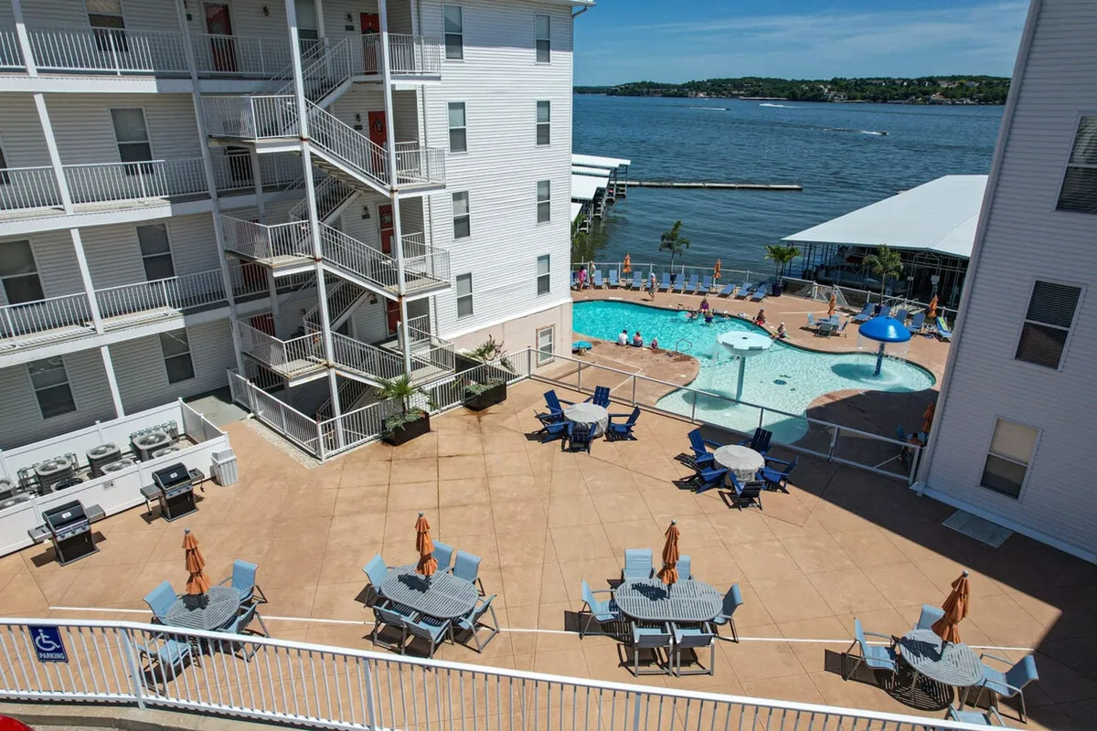A cheerful lakeside scene featuring a swimming pool, barbecue area complete with grills, lounge chairs, and an unobstructed view of the water beyond. The setting is made for outdoor meals, sunbathing, and group enjoyment.