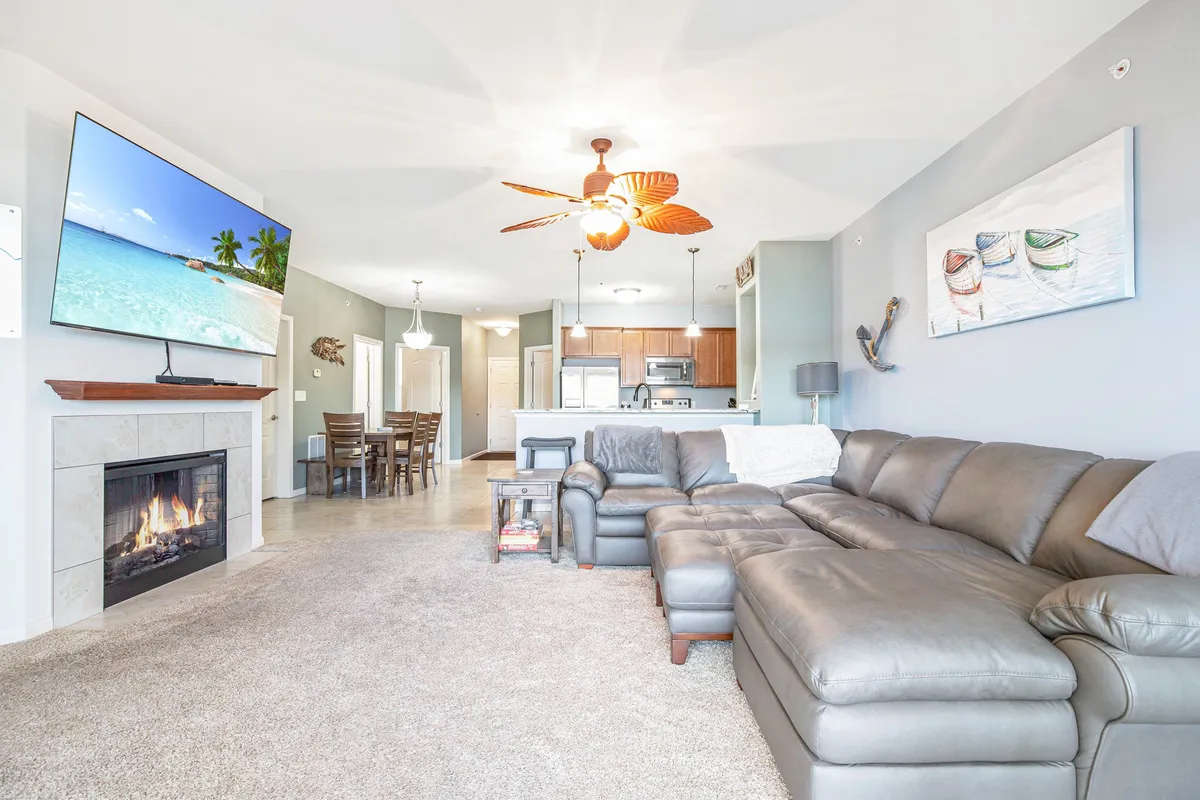 A welcoming open-concept living space featuring a comfortable leather sectional sofa arranged around a modern fireplace, with a large flat-screen TV displaying a beach scene. The dining area and kitchen are visible in the background, seamlessly connecting the main areas for socializing and family gatherings.