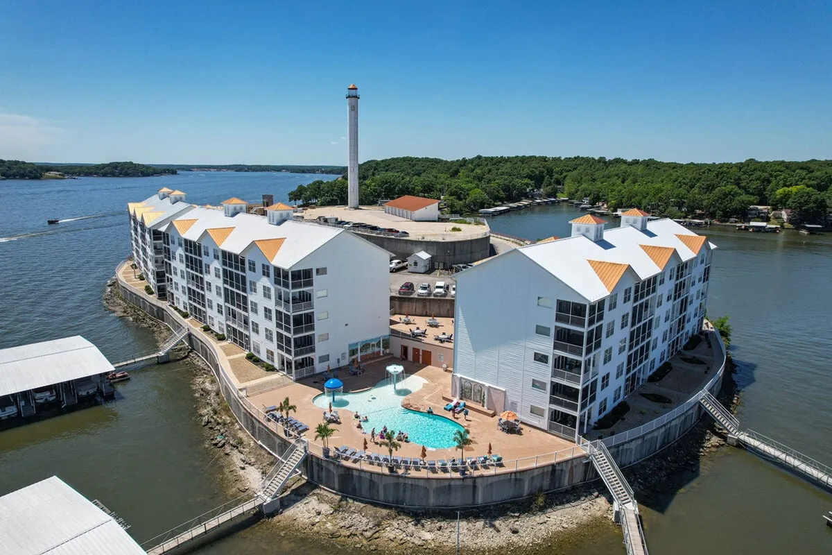 The image showcases a serene exterior view of the condo complex set on its own island, with a sparkling pool area surrounded by landscaped gardens, a unique lighthouse, and panoramic lake views. The water laps around the property, creating a private oasis atmosphere ideal for relaxation, swimming, or lakeside adventures. Sunset hues bathe the scene, hinting at tranquil evenings by the pool or atop the lighthouse.