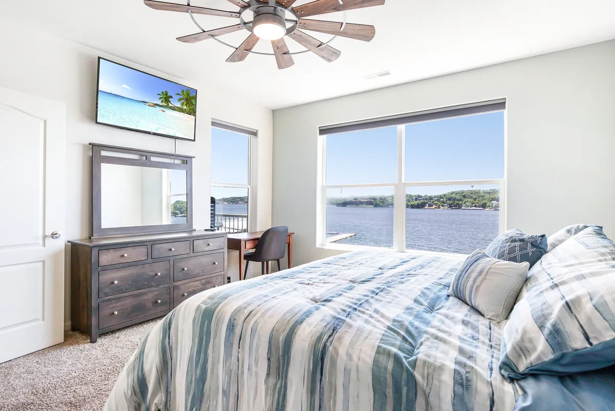 A tranquil bedroom with a large bed dressed in striped bedding, a dresser and mirror, TV mounted on the wall, and expansive windows framing a lakeside view. The space is bathed in natural light and has a ceiling fan for comfort.
