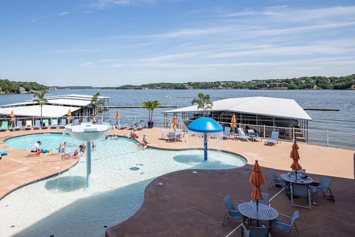 This image showcases the resort-style zero-entry swimming pool with a water fountain, surrounded by beach chairs and umbrellas, all with a sweeping view of the lake in the background. The atmosphere is lively, with people swimming, sunbathing, and socializing, suggesting both relaxation and fun.